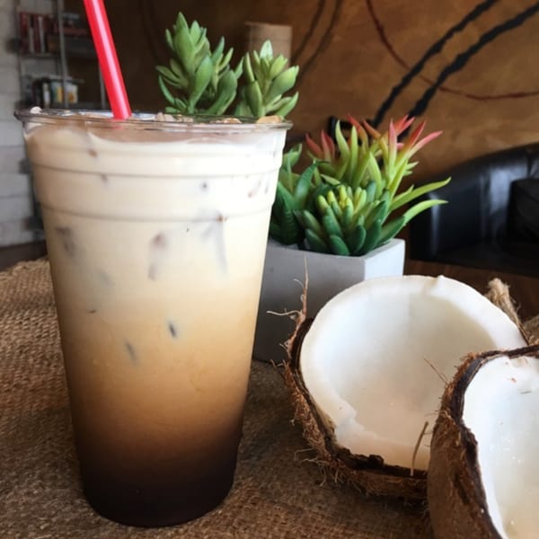 Iced coffee with a red straw, two potted succulents, and two coconut halves on a burlap surface.