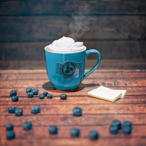 Steaming World Blend Coffee mug with whipped cream, blueberries, and white chocolate on a wooden table.