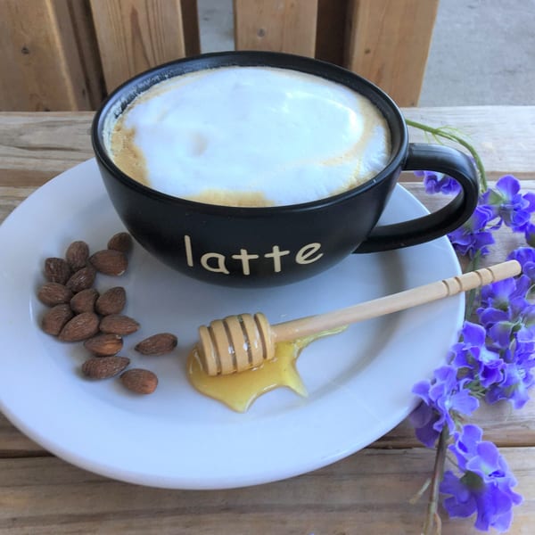 A black latte mug with foam, almonds, honey dipper with honey, and purple flowers on a white plate.