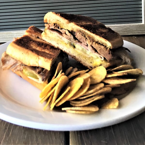 A Cuban sandwich, cut in half, with a side of plantain chips on a white plate.